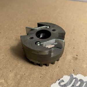 May include: A metallic gray, circular mechanical component with a toothed edge and two curved metal pieces. The component has a central hole and is secured with small screws. The background is a light brown surface.