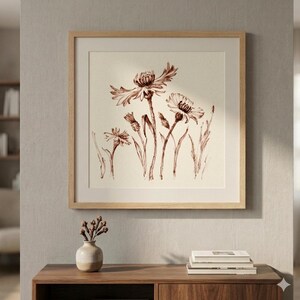 May include: A square framed wall art piece featuring a brown ink botanical illustration of wildflowers on a cream background. The artwork is enclosed in a light-toned wooden frame with a wide mat, displaying detailed sketches of several flower heads.