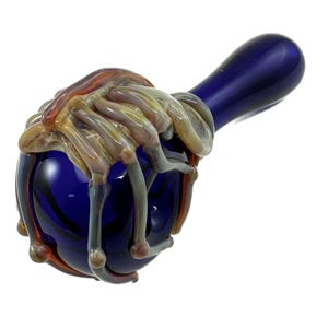 May include: A dark blue glass pipe with a bulbous bowl and a long, rounded stem. The bowl is adorned with a detailed, multi-colored glass sculpture resembling a skeletal hand. The hand sculpture features shades of orange, yellow, and gray.