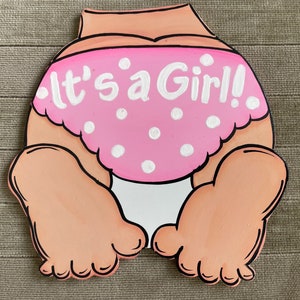 May include: A pink and white baby diaper with white polka dots and the text "It's a Girl!" painted on it. The diaper is attached to a wooden cutout of a baby's bottom.
