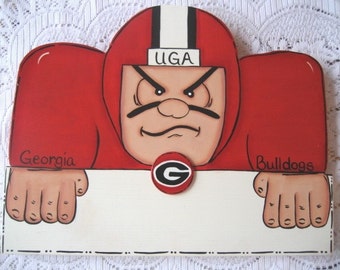 May include: A wooden sign featuring a cartoon football player wearing a red and white uniform with the word "UGA" on the helmet. The player is holding a sign with the words "Georgia Bulldogs" and a red and black "G" logo.
