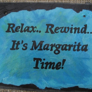 May include: A blue slate sign with white and green accents. The sign reads "Relax. Rewind. It's Margarita Time!"