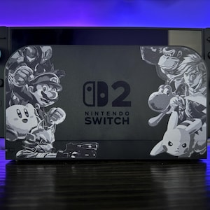 May include: Black Nintendo Switch console with a custom skin featuring Mario, Kirby, Donkey Kong, Link, Yoshi, and Pikachu. The skin has a black and white design. The console is on a dark surface.