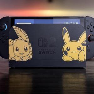 Könnte beinhalten: Eine schwarze Nintendo Switch-Konsole mit einer Dockingstation mit goldenen Evoli- und Pikachu-Designs. Die Konsole hat blaue und rote Controller. Das Dock hat das Nintendo Switch-Logo und Text. Der Bildschirm zeigt ein Menü.