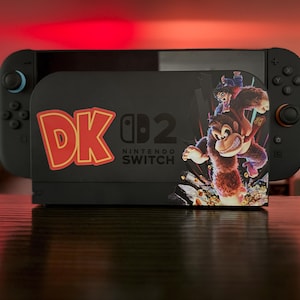 May include: A black Nintendo Switch console with a black docking station featuring a Donkey Kong design. The docking station has the text "DK" in red and yellow, and "Nintendo Switch" in white.