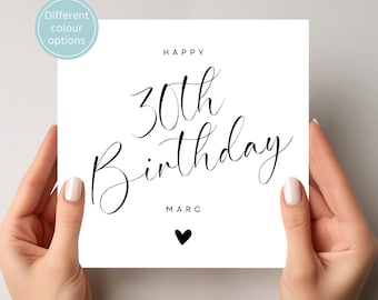 May include: A white birthday card with the words "Happy 30th Birthday Marc" in elegant black script. A small black heart is below the name. The card is held by two hands.