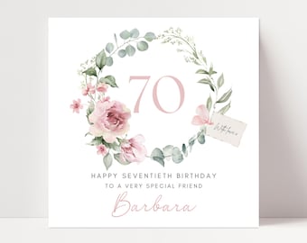 May include: A white birthday card with a floral wreath design. The card features the number 70 in a soft pink hue, surrounded by pink flowers and green foliage. Text includes "Happy Seventieth Birthday" and a name.