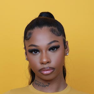 May include: A portrait of a person with dark skin and long eyelashes against a yellow backdrop. The person has their hair up in a bun. They are wearing a mustard-coloured top and have a tattoo on their neck.