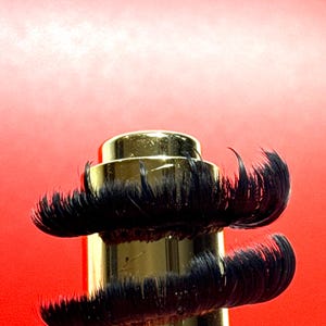 Könnte beinhalten: Ein goldfarbenes, zylindrisches Objekt mit zwei Schichten aus schwarzen, gebogenen, wimpernartigen Verlängerungen. Der Hintergrund ist ein Farbverlauf aus Rottönen.