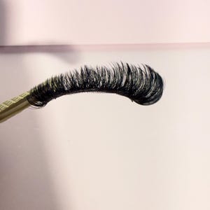 Könnte beinhalten: Ein einzelnes, gebogenes, schwarzes falsches Wimpernpaar, das von einer silbernen Pinzette gehalten wird. Die Wimper ist dicht mit einzelnen, dunklen Fasern gefüllt, was ein volles, voluminöses Aussehen erzeugt. Der Hintergrund ist ein weiches, helles Rosa.