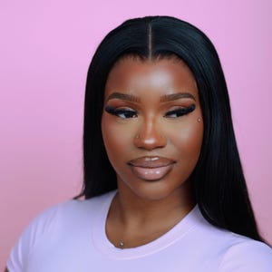 Op de afbeelding: Een persoon met lang, steil zwart haar en volledige make-up, inclusief donkere wimpers en lipgloss, wordt getoond tegen een roze achtergrond. De persoon draagt een lichtpaars t-shirt en heeft een neusring en een kleine piercing onder de lip.