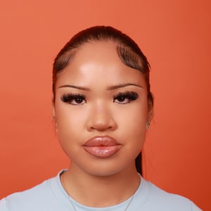 May include: A portrait of a person with dark hair and styled baby hairs. The person is wearing a light blue top and a delicate necklace. The background is a solid orange color. The person has full lips and long eyelashes.