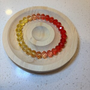 May include: A bracelet featuring faceted beads in a colour gradient from yellow to red. The bracelet is displayed on a small, circular wooden dish. The beads transition from yellow to orange to red.