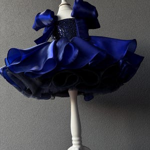 May include: A striking blue dress with a sequined bodice and a layered skirt. The dress has puffed sleeves and is displayed on a white mannequin. The skirt is made of multiple layers of ruffled fabric.