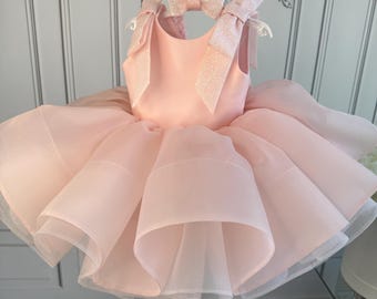 May include: A light pink tutu dress with a matching bow and shoulder straps. The dress is hanging on a white hanger. The bow and straps have a glittery finish. The skirt is layered with tulle.