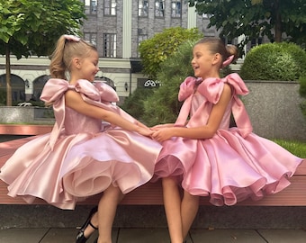 May include: Two matching sleeveless dresses in a dusty rose pink hue, featuring voluminous layered skirts with ruffled hems and large, prominent bows tied at each shoulder. The bodice is fitted, and the fabric has a subtle, shimmering finish.