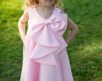 May include: A sleeveless light pink dress featuring a large, structured bow detail on the front bodice and a layered tulle hemline. The dress has a V-neckline and a smooth, satin-like finish, paired with simple white flat shoes and a pearl headband.