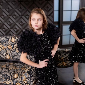 Peut inclure: Une jeune personne porte une robe noire à paillettes avec des détails d'épaules en plumes. La robe arrive au genou et a un col rond. L'arrière-plan présente un mur à motifs et un canapé à motifs floraux.
