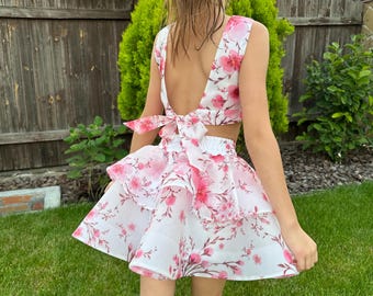 May include: A two-piece floral outfit featuring a crop top and tiered skirt. The white fabric is adorned with pink floral patterns. The top has a low back with a bow. The skirt has multiple layers of ruffled fabric. The outfit is designed for a child.