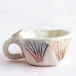 Puede incluir: Taza de cerámica hecha a mano, de forma irregular. La taza blanquecina tiene un asa y está decorada con patrones verticales en forma de abanico, en tonos rojos, azules y verdes. Una pieza de arte única, ideal para café o té.