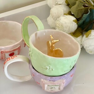May include: A stack of pastel-coloured ceramic mugs. The top mug is light green with a gold handle and a gold bunny figurine inside. The bottom mug is lavender with the text "Matcha Girl".