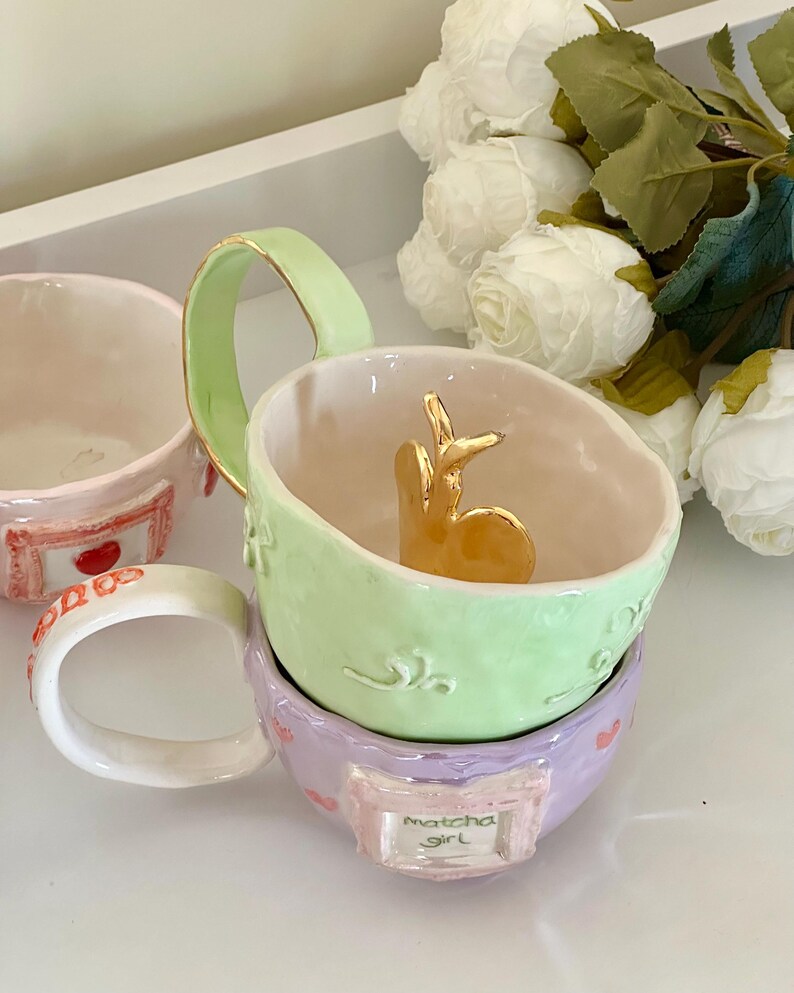 May include: A stack of pastel-coloured ceramic mugs. The top mug is light green with a gold handle and a gold bunny figurine inside. The bottom mug is lavender with the text "Matcha Girl".