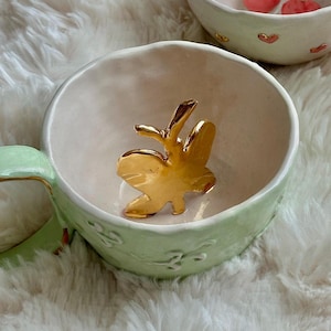 May include: A pale green ceramic mug featuring a gold butterfly figurine inside. The mug has a handle and embossed floral patterns. Another bowl with cherries is visible in the background. The mug is on a white, fluffy surface.