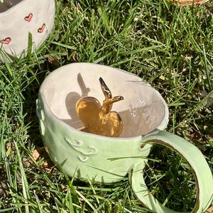 May include: A pale green ceramic mug with a gold apple sculpture inside. The mug has a rounded handle and decorative swirls. Other ceramic mugs are visible in the background, one with pink heart designs.