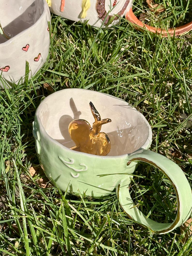 May include: A pale green ceramic mug with a gold apple sculpture inside. The mug has a rounded handle and decorative swirls. Other ceramic mugs are visible in the background, one with pink heart designs.