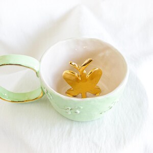 May include: A mint green ceramic teacup with a gold rim and handle. Inside the cup is a gold butterfly figurine. The cup has a white interior and a floral design on the exterior. The teacup is on a white surface.