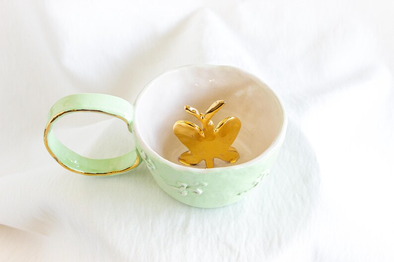 May include: A mint green ceramic teacup with a gold rim and handle. Inside the cup is a gold butterfly figurine. The cup has a white interior and a floral design on the exterior. The teacup is on a white surface.