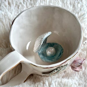 Könnte beinhalten: Eine weiße Keramiktasse mit goldfarbenem Rand und Henkel. Im Inneren der Tasse befindet sich am Boden eine modellierte blau-weiße Muschel mit einer kleinen, runden, schimmernden weißen Perle. Eine kleine rosa Muschel ist an der Außenseite angebracht.