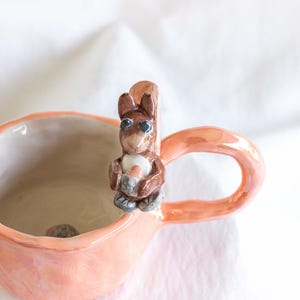 Könnte beinhalten: Pfirsichfarbene Keramiktasse mit einer Eichhörnchenfigur am Henkel. Das Eichhörnchen ist braun und grau und hält eine Nuss. Die Tasse hat eine glänzende Oberfläche und steht vor einem weißen Hintergrund.