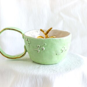 May include: A pale green ceramic mug with a gold-edged handle and rim. The mug has embossed floral patterns and a gold accent inside. A gold decorative element is inside the mug.