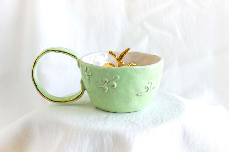 May include: A pale green ceramic mug with a gold-edged handle and rim. The mug has embossed floral patterns and a gold accent inside. A gold decorative element is inside the mug.