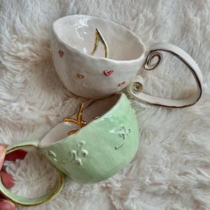 May include: Two ceramic mugs. One is white with pink hearts and gold accents, the other is light green with gold trim and embossed floral patterns. Both have distinctive handles and interior details. The mugs are handmade.