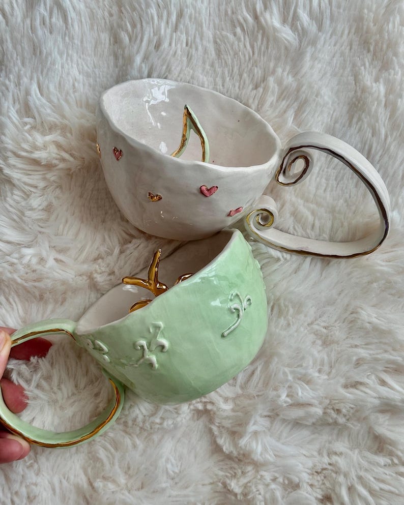 May include: Two ceramic mugs. One is white with pink hearts and gold accents, the other is light green with gold trim and embossed floral patterns. Both have distinctive handles and interior details. The mugs are handmade.