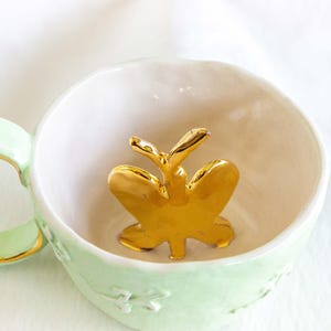 May include: A light green ceramic teacup with a gold-coloured rim and handle. Inside the cup is a gold butterfly figurine. The cup has a white interior and a floral design on the exterior.