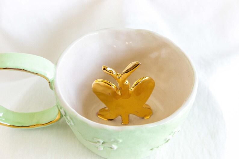 May include: A light green ceramic teacup with a gold-coloured rim and handle. Inside the cup is a gold butterfly figurine. The cup has a white interior and a floral design on the exterior.