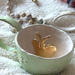 May include: A pale green ceramic cup with a gold heart-shaped ornament inside. The cup features a handle and floral and swirl patterns. A pearl necklace, a makeup brush, and a white ring with gold detailing are also present.