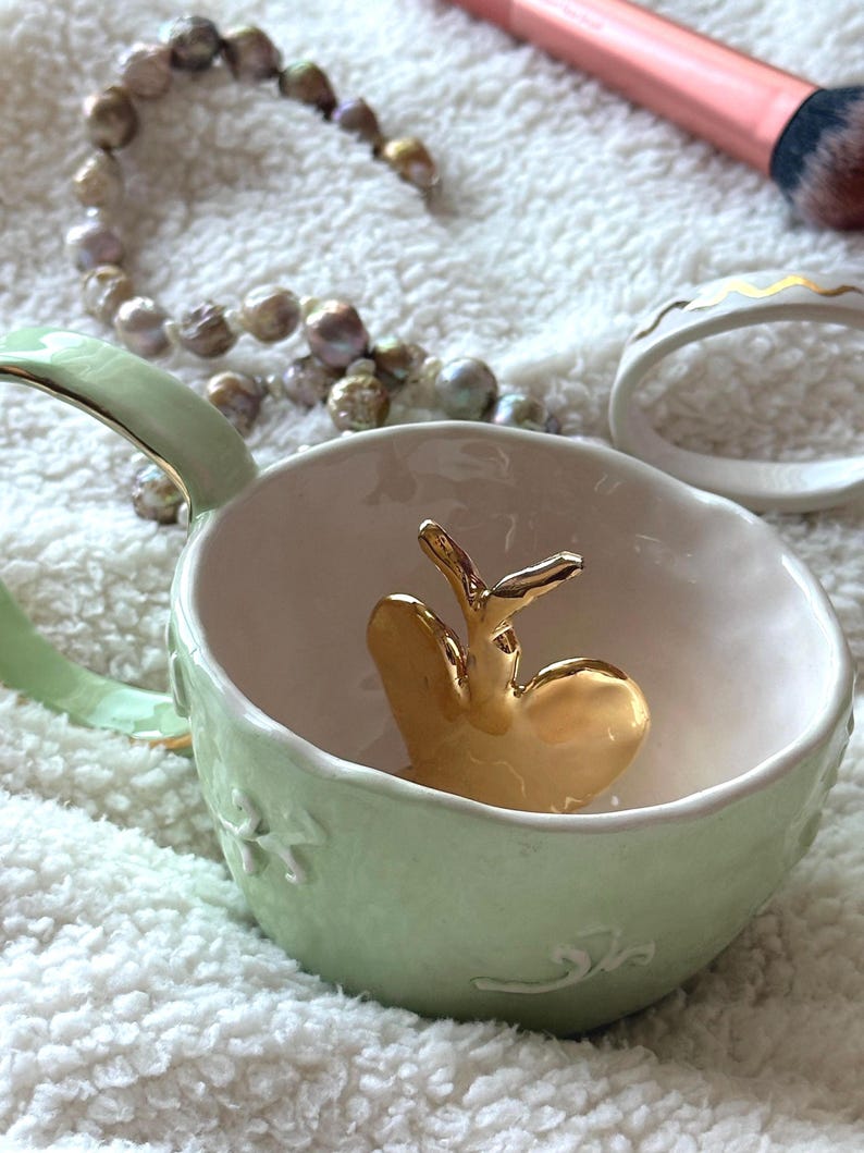 May include: A pale green ceramic cup with a gold heart-shaped ornament inside. The cup features a handle and floral and swirl patterns. A pearl necklace, a makeup brush, and a white ring with gold detailing are also present.
