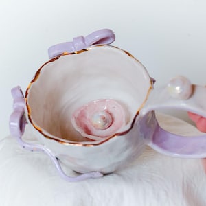 Puede incluir: Una taza de cerámica hecha a mano con un diseño único. La taza presenta un exterior lila claro con ribete dorado y un interior rosa delicado. Un pequeño lazo decorativo y un asa se suman al encanto de la taza. La taza es sostenida por una mano.