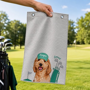 Puede incluir: Una toalla de golf de color gris claro con textura de gofre que presenta una ilustración impresa de un perro dorado con una gorra verde etiquetada como Harper, junto a una bolsa de golf verde con el nombre Samuel en cursiva verde y una bandera.