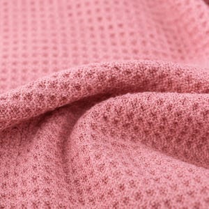 Puede incluir: Una tela de color rosa palo con una textura de tejido de gofre distintiva. El material est&aacute; doblado, creando crestas suaves y redondeadas y sombras profundas que resaltan el patr&oacute;n de cuadr&iacute;cula de la superficie textil en el encuadre.