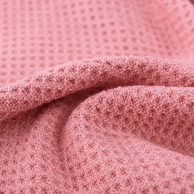 Puede incluir: Una tela de color rosa palo con una textura de tejido de gofre distintiva. El material est&aacute; doblado, creando crestas suaves y redondeadas y sombras profundas que resaltan el patr&oacute;n de cuadr&iacute;cula de la superficie textil en el encuadre.