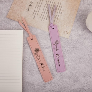 May include: Two leather bookmarks, one pink and one lavender. The pink bookmark has a floral design and the name "Alex". The lavender bookmark has a rose design and the name "Ronan". Both have a stitched border and a ribbon.