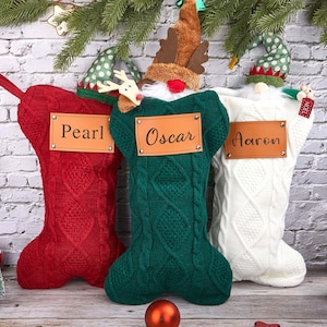 Puede incluir: Tres calcetines navideños en rojo, verde y blanco, cada uno con un diseño de punto trenzado. El calcetín rojo tiene "Pearl" en una etiqueta de cuero, el verde "Oscar" y el blanco "Aaron". Cada calcetín está decorado con un personaje festivo.