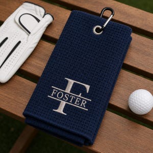 May include: A navy blue waffle-textured golf towel featuring an embroidered silver letter F and the name FOSTER. The towel includes a silver metal grommet and a black carabiner clip. A white golf glove and a white golf ball are positioned nearby.