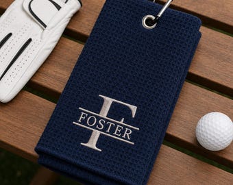 Puede incluir: Una toalla de golf azul marino con textura de gofre que presenta una letra F plateada bordada y el nombre FOSTER. La toalla incluye un ojal de metal plateado y un mosquetón negro. Un guante de golf blanco y una pelota de golf están cerca.