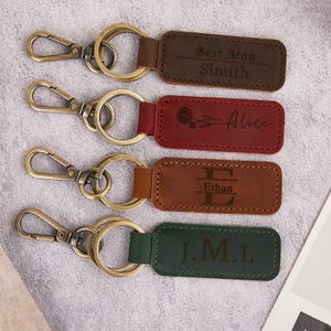 May include: Four personalised leather keyrings with metal clasps. The keyrings are brown, red, tan, and green. Each keyring has a different name or initials engraved on it. The first keyring says "Best Man Simith". The second keyring says "Alice". The third keyring says "Ethan". The fourth keyring says "J.M.L".